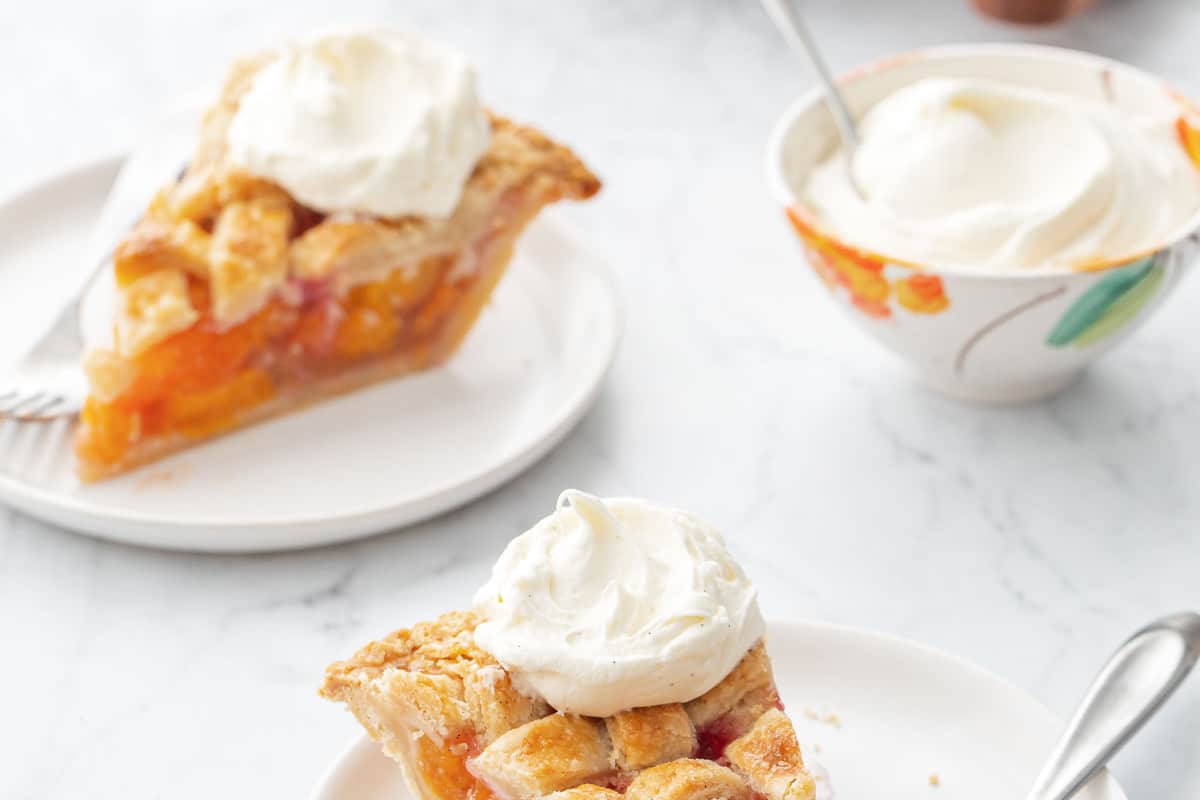 Slices of Vanilla Bean Peach Pie on white plates with forks and topped with dollops of whipped cream, with a small bowl of cream and the rest of the pie in the background.