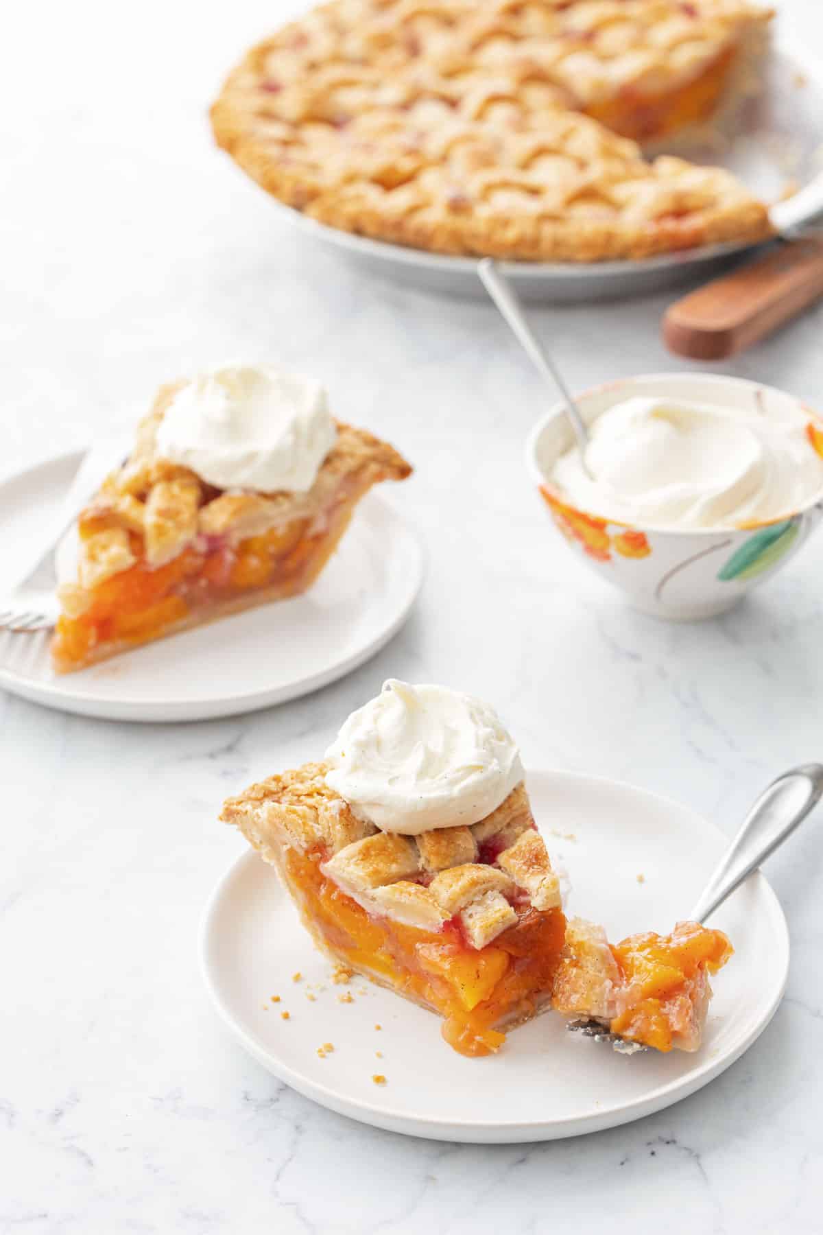 Slices of Vanilla Bean Peach Pie on white plates with forks and topped with dollops of whipped cream, with a small bowl of cream and the rest of the pie in the background.