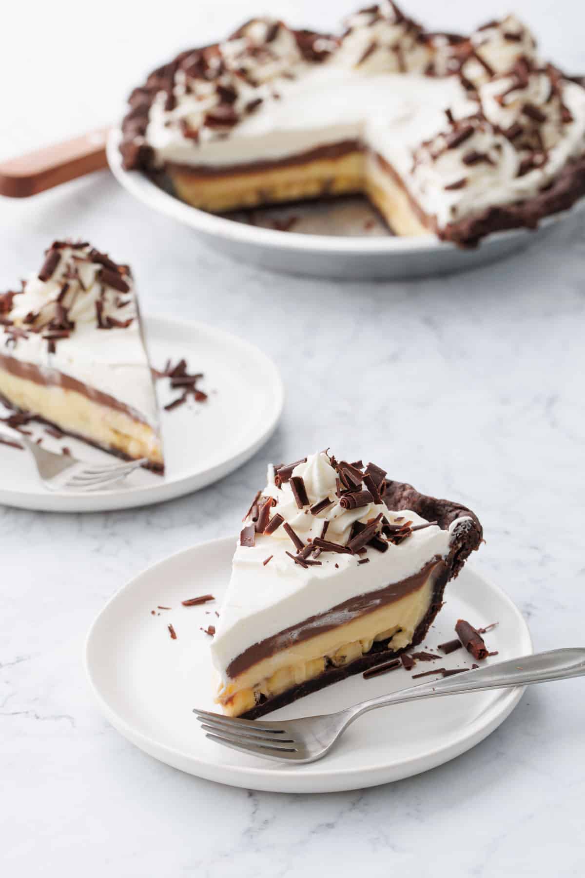 Chocolate Banana Cream Pie with Flaky Chocolate Pie Crust sliced on white plates on marble background.