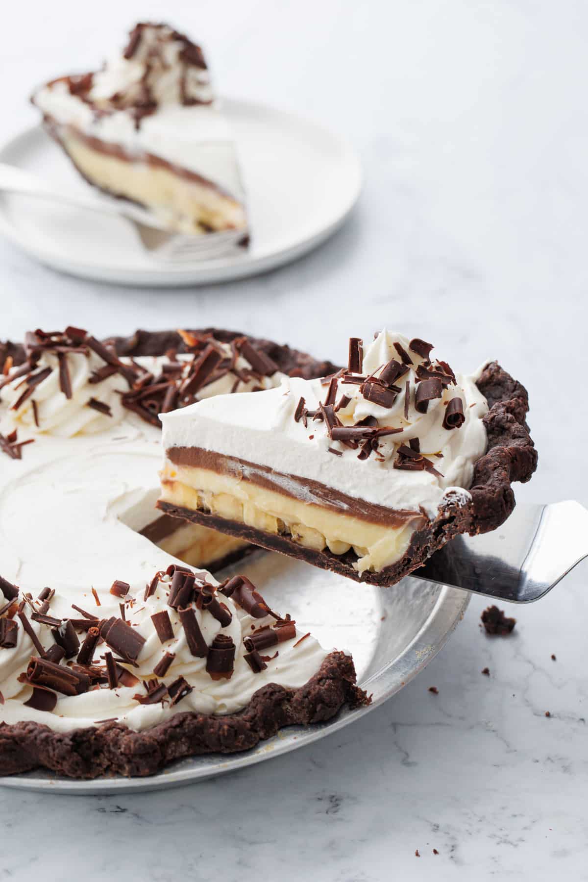Lifting a slice of Chocolate Banana Cream Pie out of a pie plate on a white marble background.