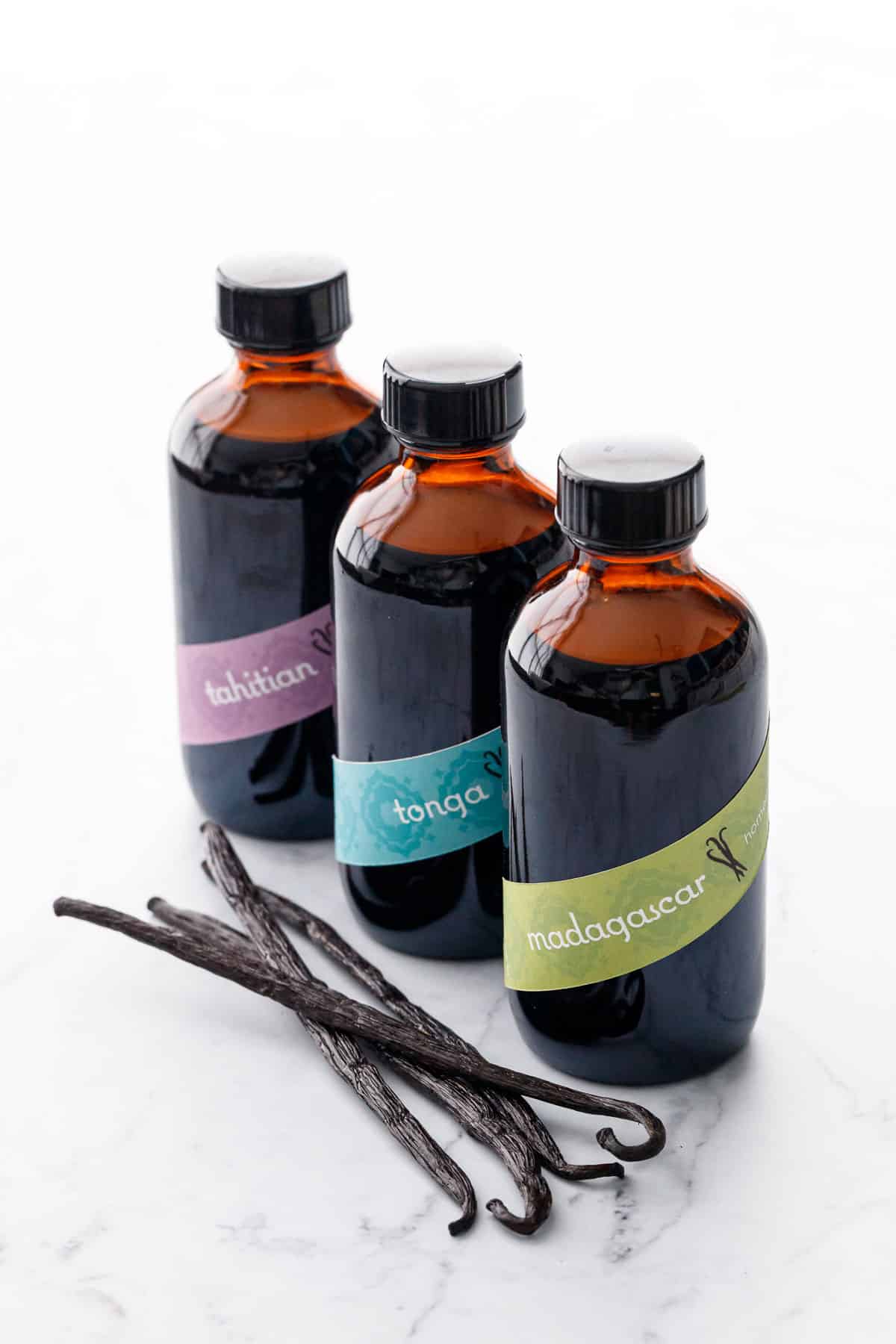 Row of three amber glass bottles filled with Homemade Vanilla Extract, with colorful diagonal band labels and a few vanilla beans on the side.