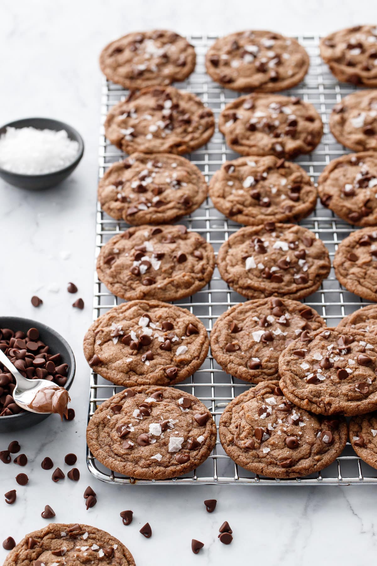Rows of Nutella Chocolate Chip Cookies on a wire baking rack, with bowls of chocolate chips, flake salt, and a spoonful of Nutella on the side.