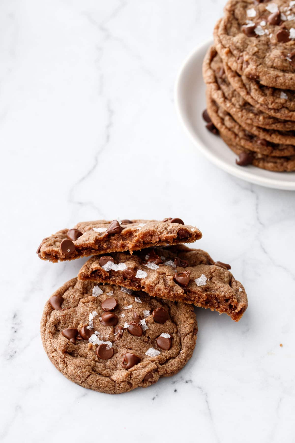 Two Nutella Chocolate Chip Cookies, one broken in half to show the interior texture, on a marble background with a plate of more cookies in the back.