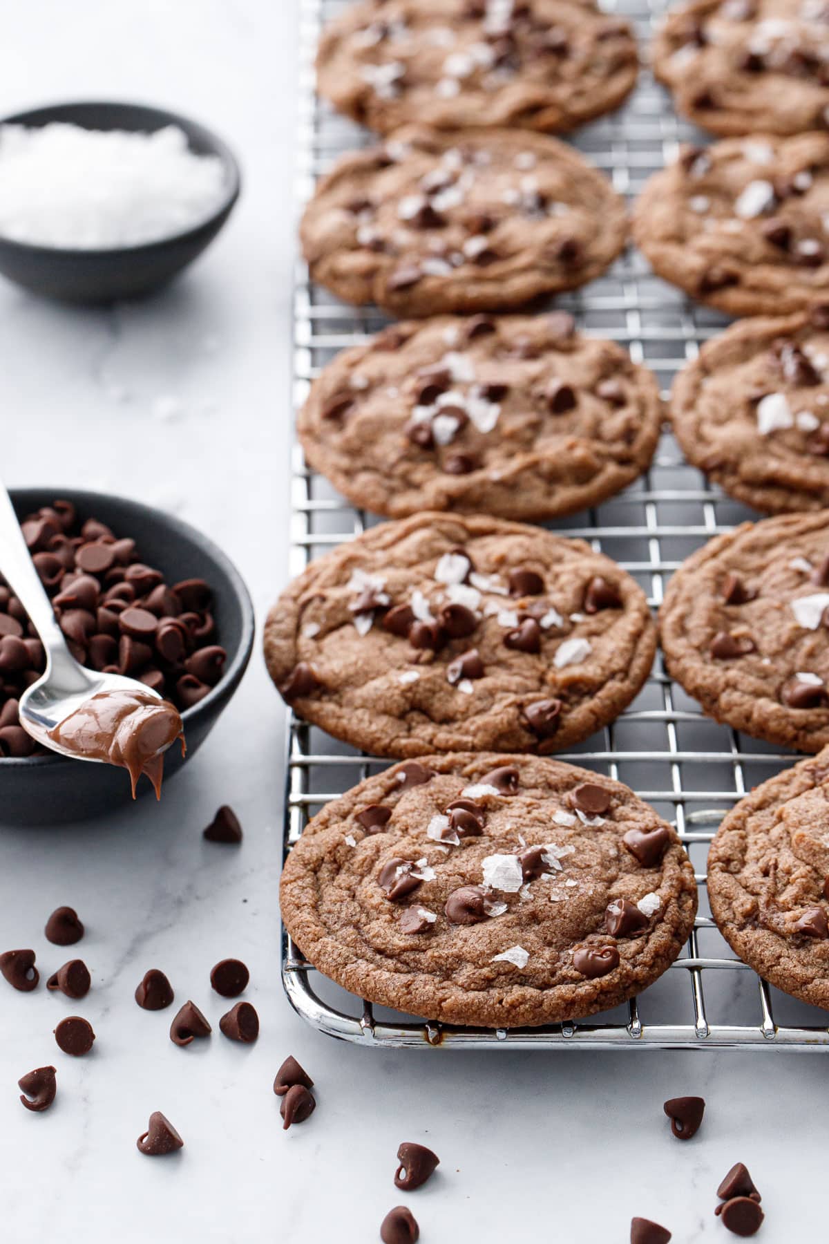 Rows of Nutella Chocolate Chip Cookies on a wire baking rack, with bowls of chocolate chips, flake salt, and a spoonful of Nutella on the side.
