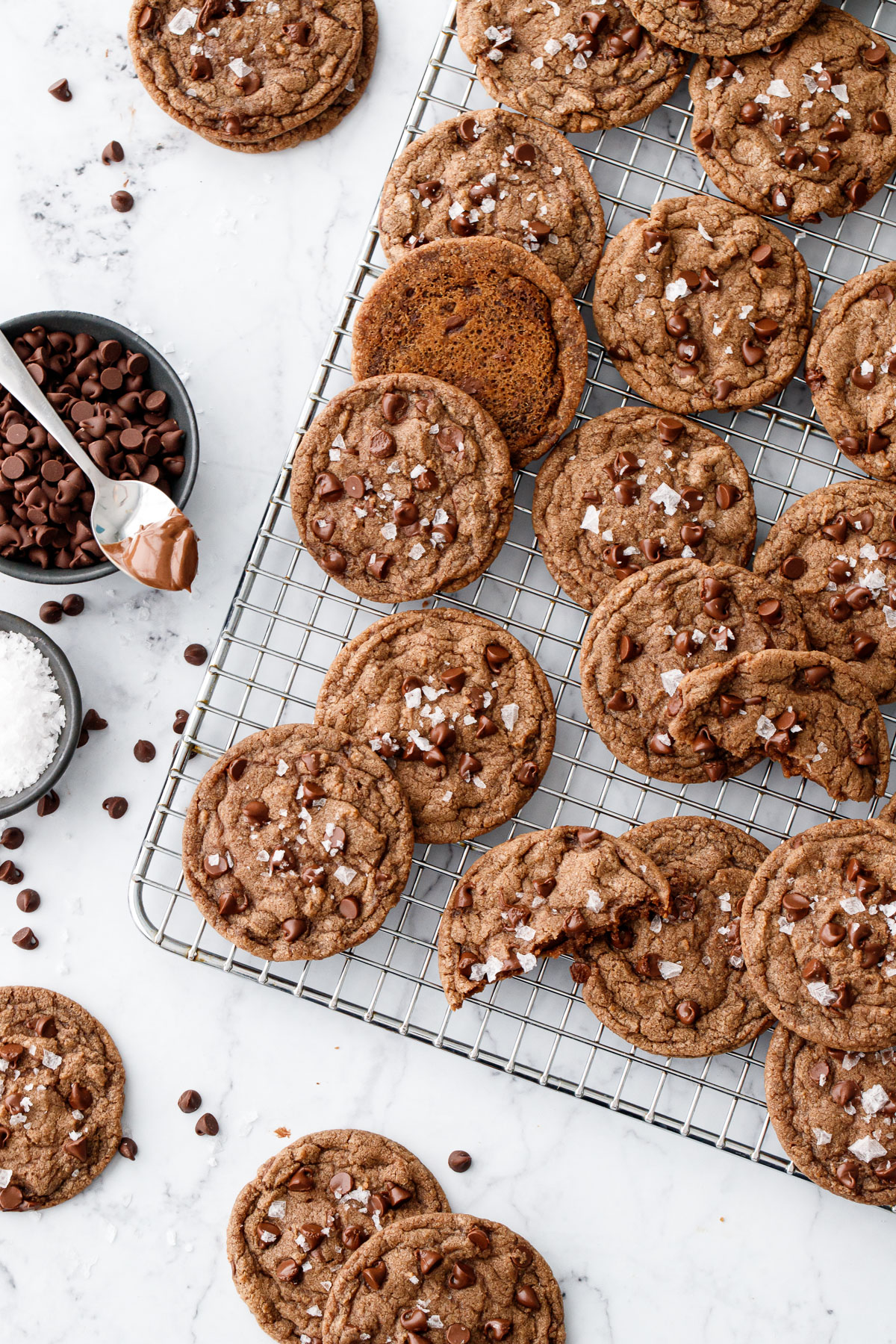 Scattered Nutella Chocolate Chip Cookies on a wire baking rack, with bowls of chocolate chips, flake salt, and a spoonful of Nutella on the side.