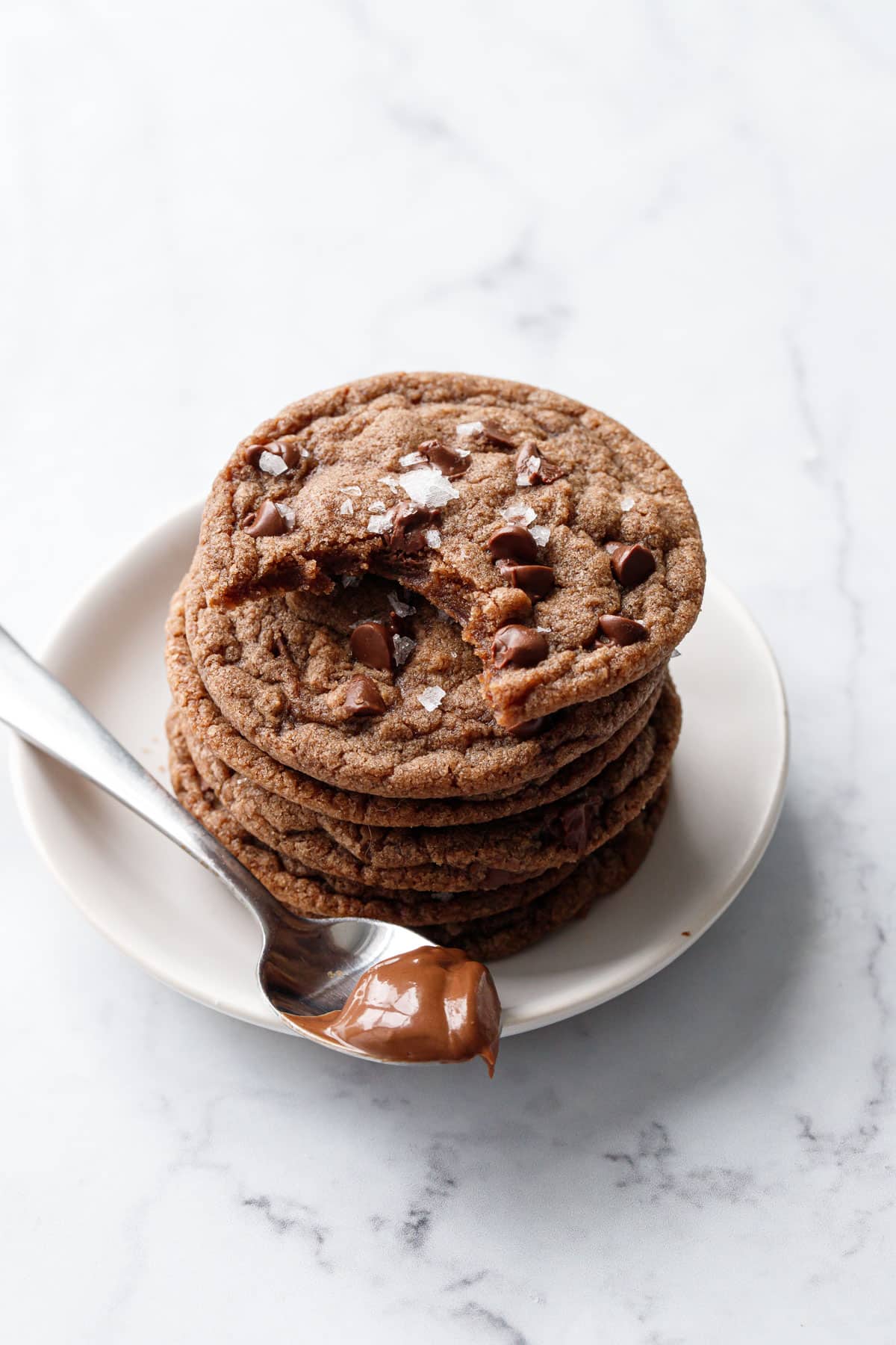 Stack of Nutella Chocolate Chip Cookies, one cookie with a bite out of it, and a spoonful of Nutella on the side of the plate.