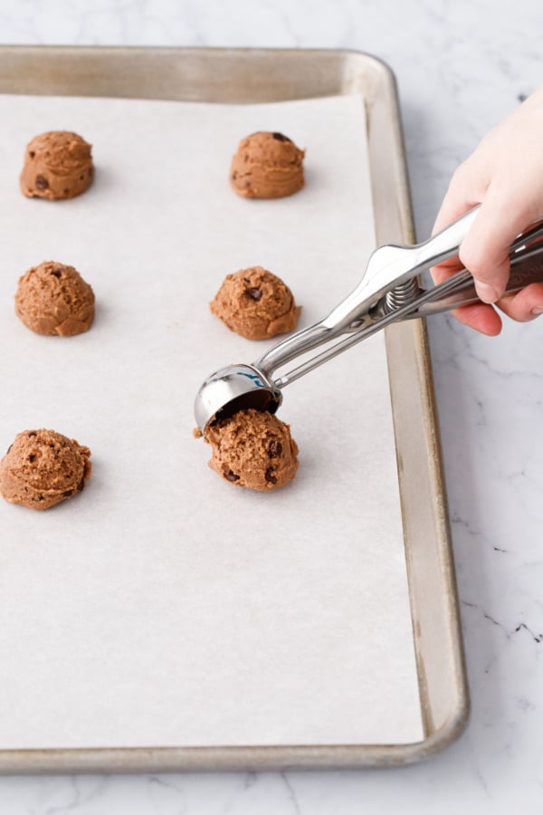 Small cookie scoop scooping small balls of cookie dough onto a parchment-lined baking sheet.