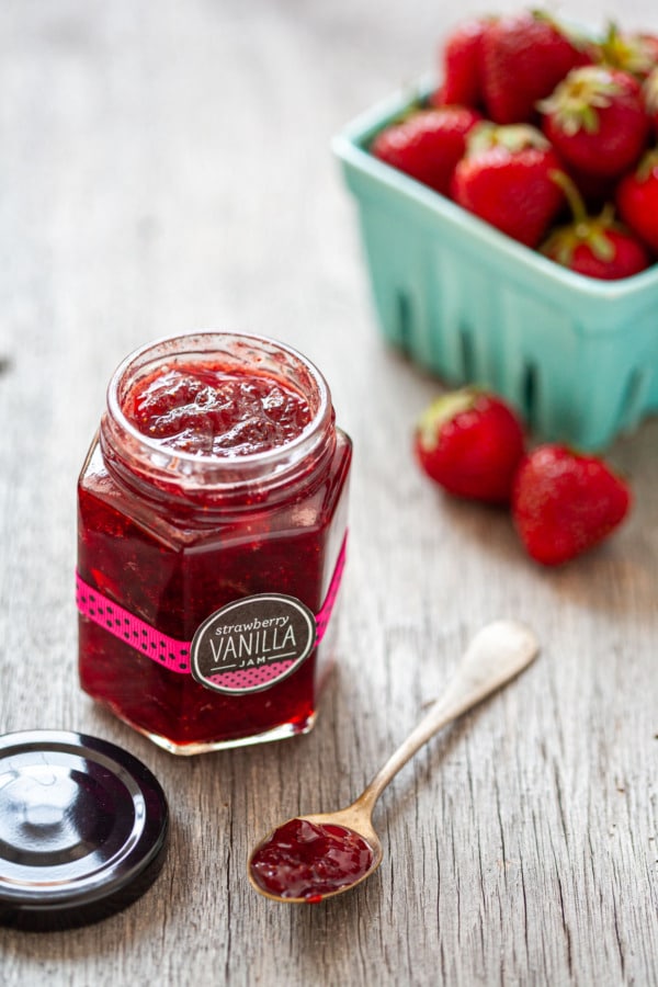 Jar of Strawberry Vanilla Jam on a rustic wood background with a pint of fresh strawberries in the background.