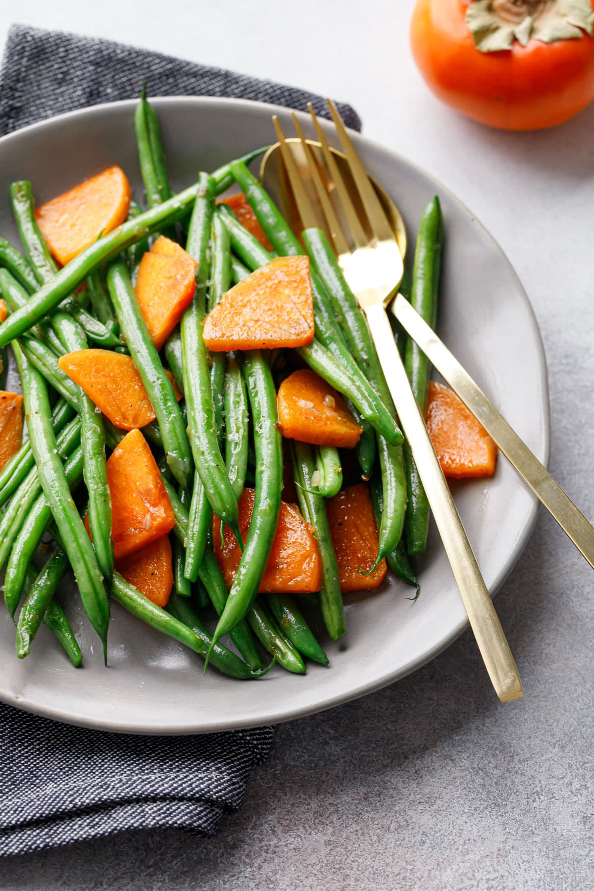 Gray plate with Sautéed Green Beans and Persimmons, with gold serving utensils one Fuyu persimmon in the background.
