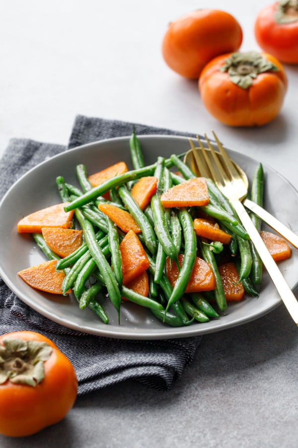 Gray serving plate wtih Sautéed Green Beans and Persimmons, gold serving utensils, and whole Fuyu persimmons in the foreground and background.