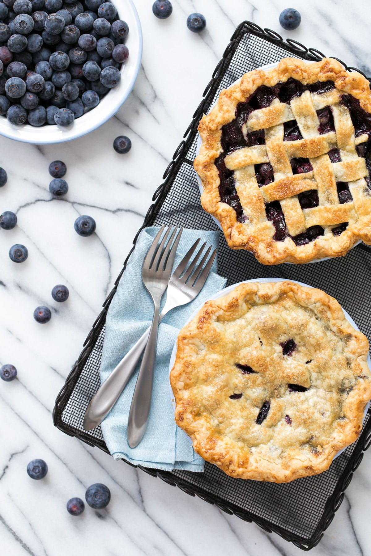 Overhead, two mini blueberry pies on a wire rack, with a bowl of berries on the side and more scattered around.