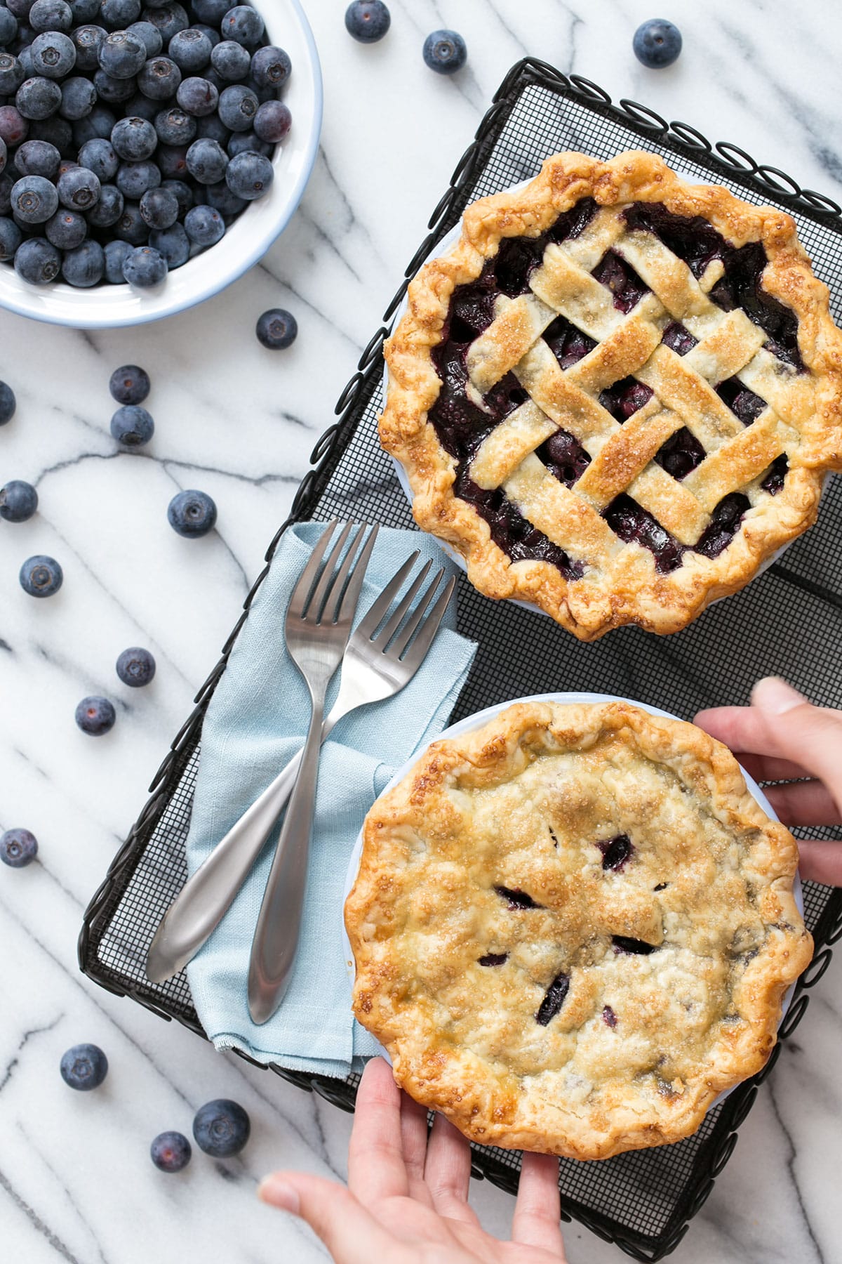 Overhead, two mini blueberry pies on a wire rack, with a bowl of berries on the side and more scattered around.