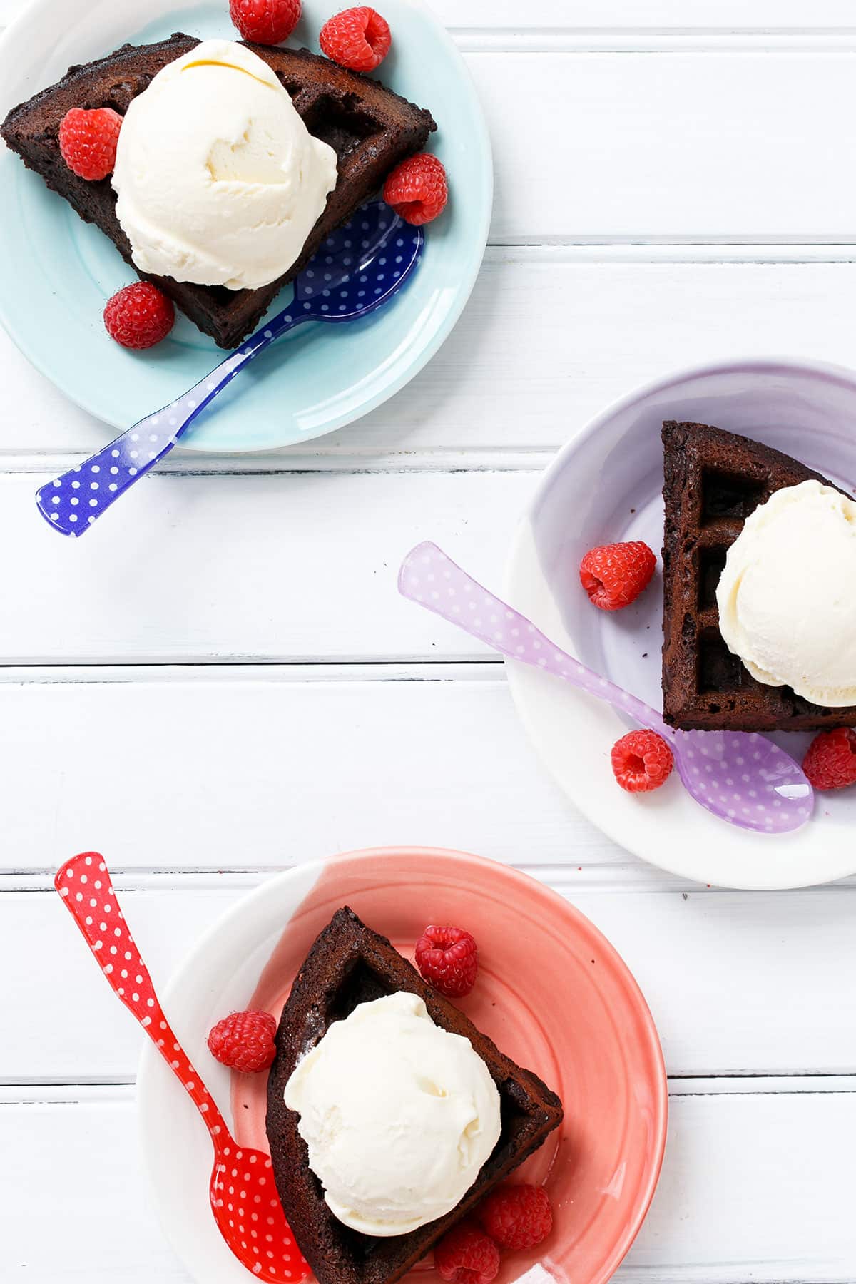 Overhead, three colorful plates each with a quarter chocolate waffle, with fresh berries and colorful plastic spoons.