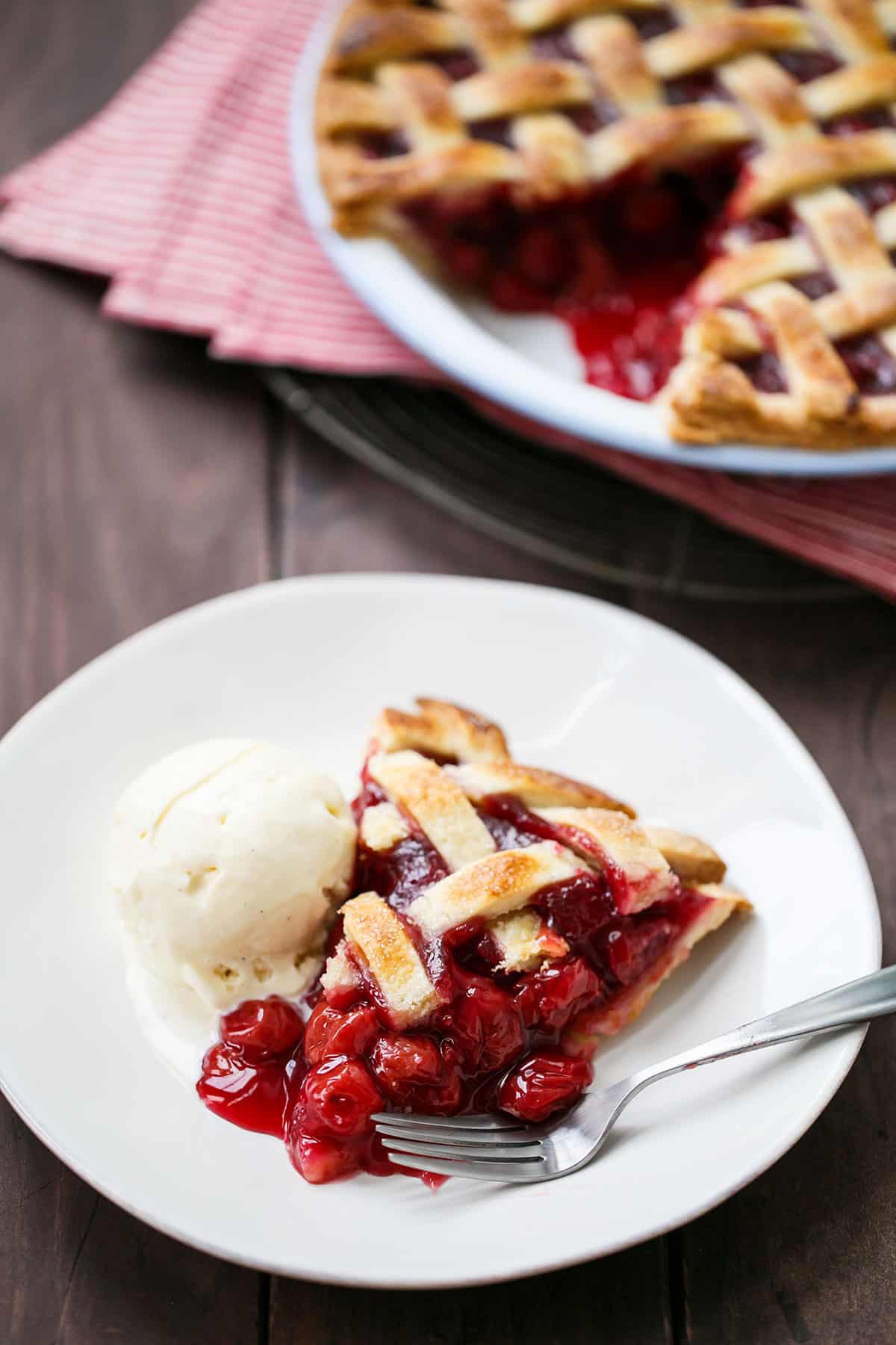 One slice of Tart Cherry Pie on a white plate with a scoop of ice cream, the full pie in the background.