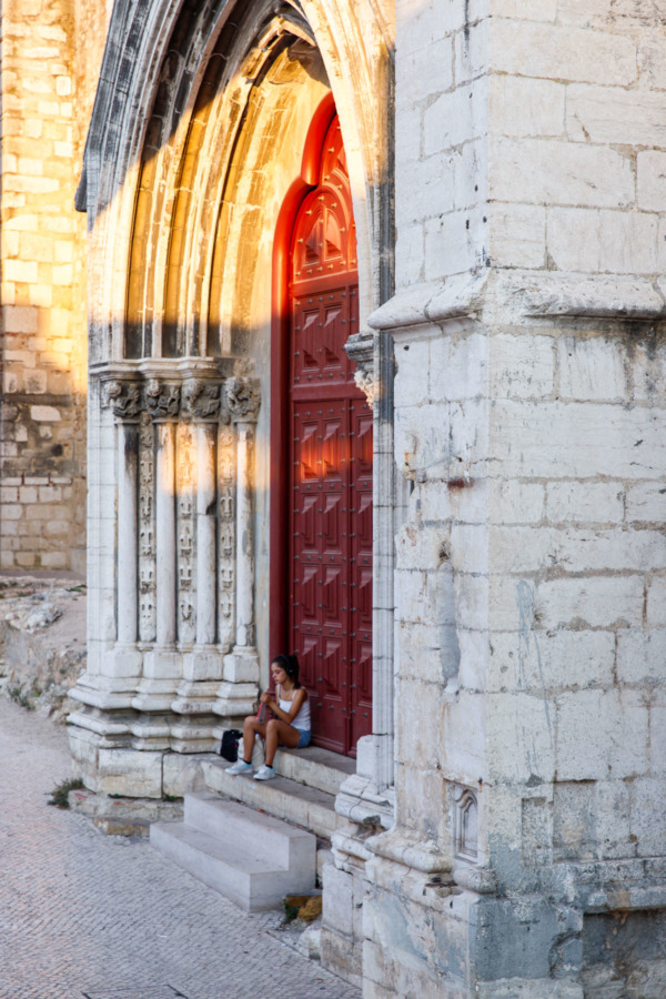 Girl sitting in front of the large red side door of the Carmo Convent in Lisbon, Portgual