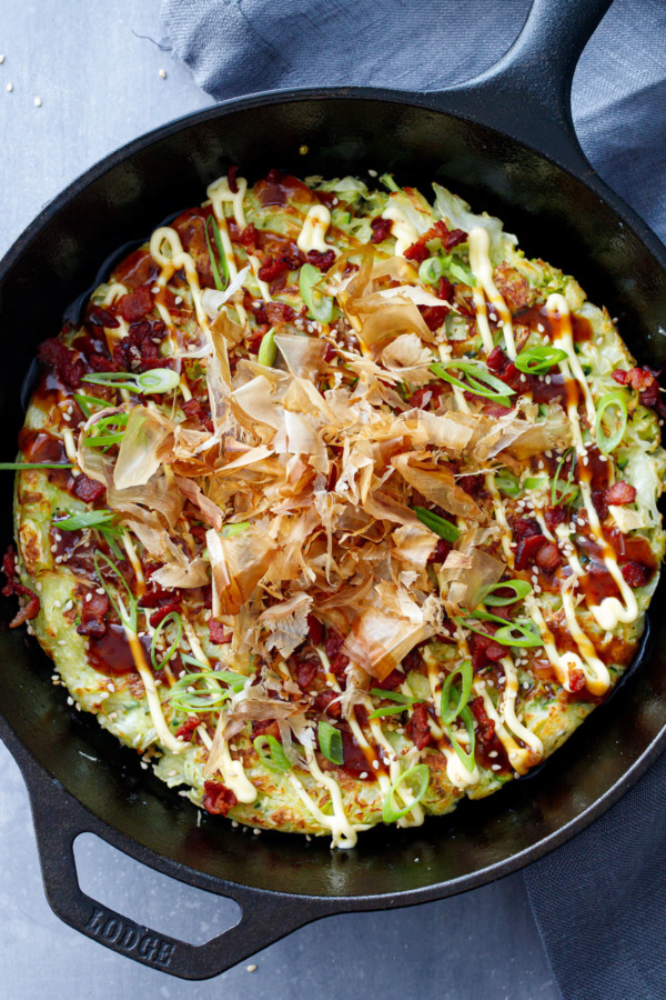 Overhead shot of cast iron skillet with okonomiyaki (Japanese Savory pancake), drizzled with sauce and topped with bonito flakes