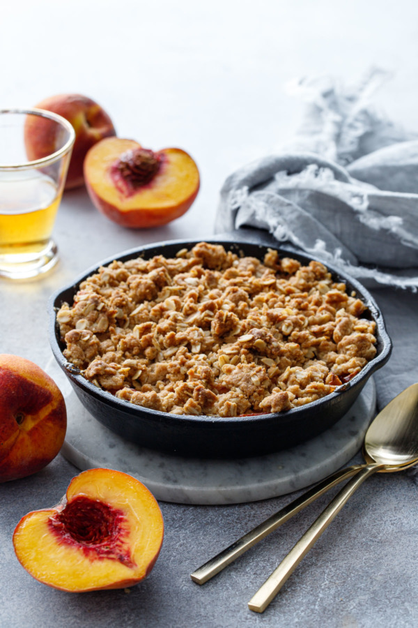 Bourbon Peach Crisp made in a cast iron skillet just out of the oven, with fresh peaches and a shot glass of bourbon in the background.