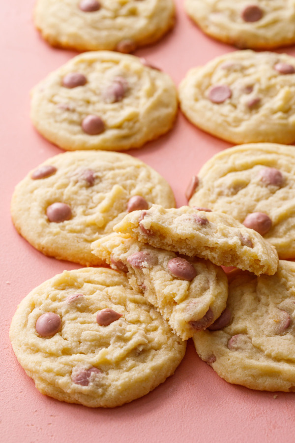 Two rows of Cacao Butter and Ruby Chocolate Chip Cookies on a pink background, with one cookie broken in half on top