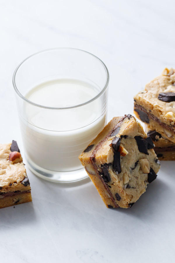 Square cut blondies on a white marble background, one leaning against a glass of milk.