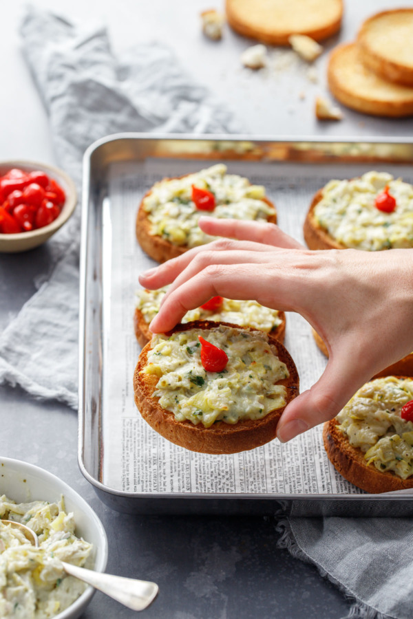 Hand lifting one artichoke crostino toast off of the baking pan