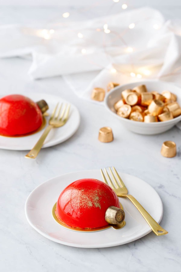 Two red mirror-glazed cakes decorated to look like Christmas ornaments, on white plates with gold forks and fairy lights in the background.