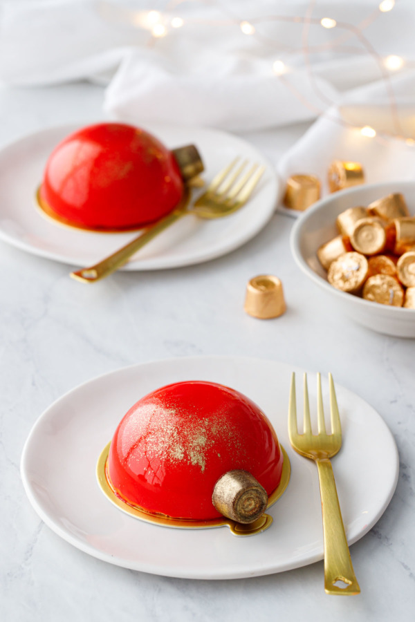 Two red mirror-glazed cakes decorated to look like Christmas ornaments, on white plates with gold forks and fairy lights in the background.