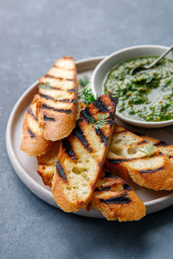 Plate of thin slices of grilled baguette bread and a bowl of carrot top pesto with a wee spoon.