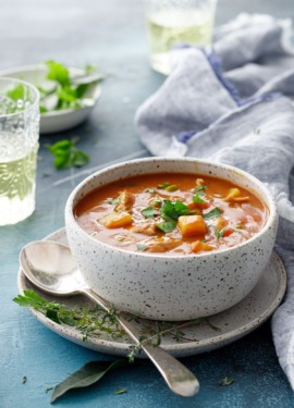 Handmade ceramic bowl filled with hearty turkey stew, on a blue background with blue napkin, wine glasses and a bit of parsley in the background.