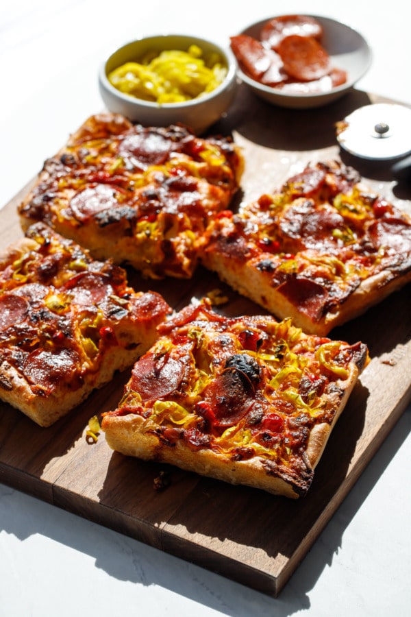 Sliced Detroit-Style Pepperoni and Pickled Pepper Pan Pizza, shot in harsh light on a wooden cutting board