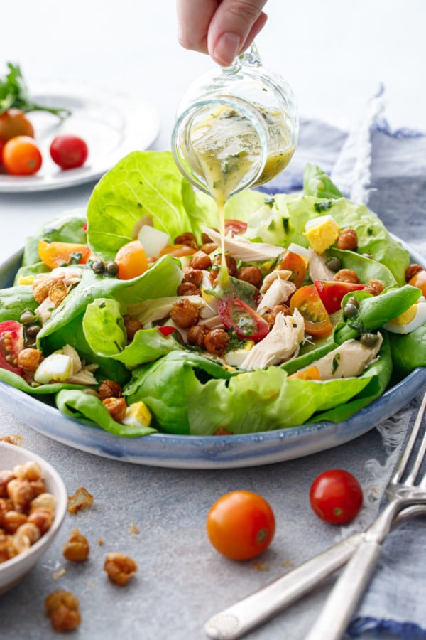 Hand pouring dressing out of a small glass pitcher onto a shallow dish with large leaves of butter lettuce and nicoise toppings.
