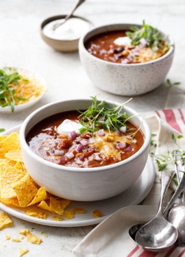 Two white bowls of Vegetarian Pumpkin & Three-Bean Chili topped with cheese, sour cream and micro cilantro