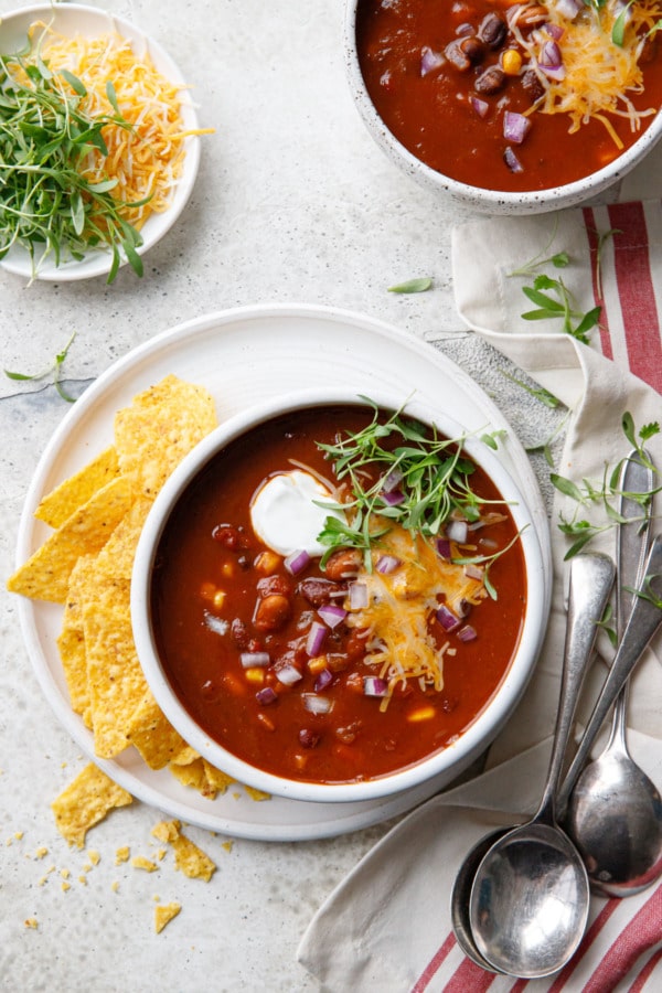Overhead, two bowls of Vegetarian Pumpkin & Three-Bean Chili with spoons and toppings