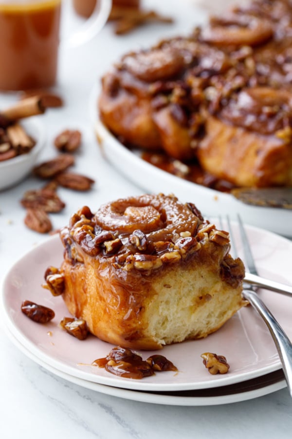 Single hot buttered rum sticky bun on a light pink plate, with the platter of buns and a cup of buttered rum in the background
