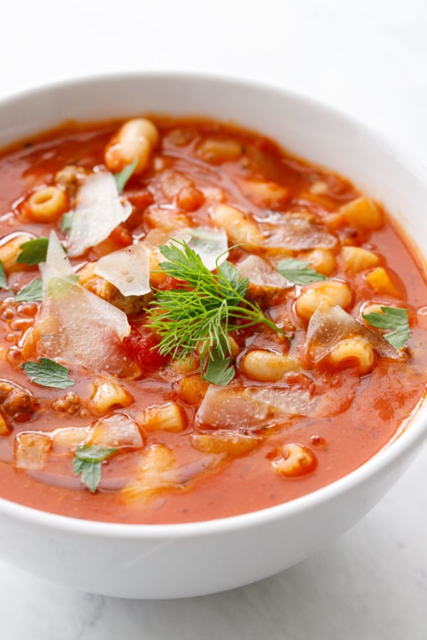 Closeup white bowl with Pasta e Fagioli soup, topped with parmesan, parsley and fennel fronds