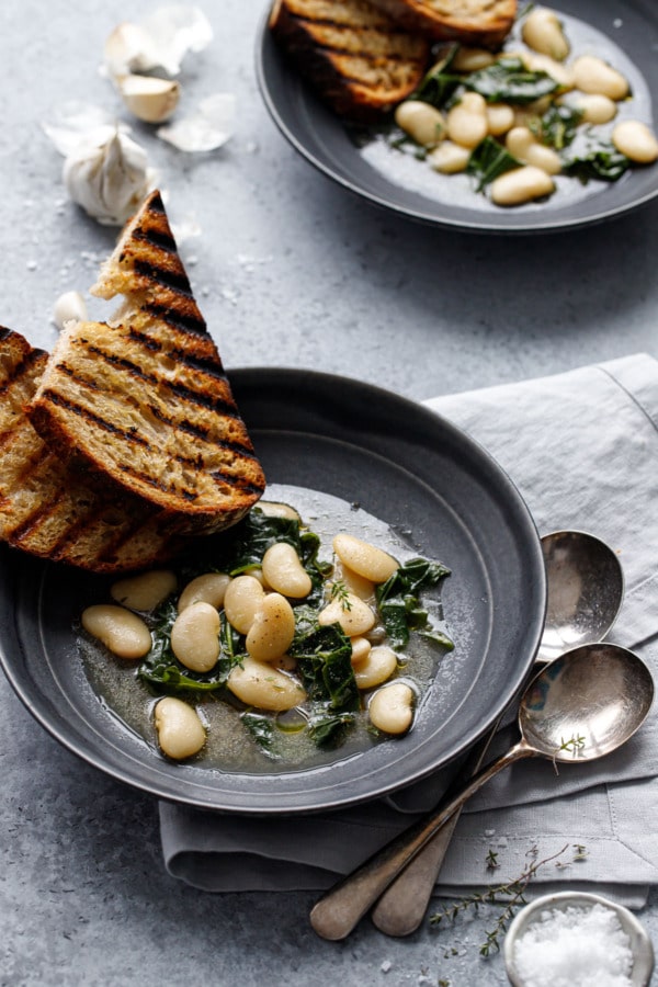 Two charcoal gray bowls with brothy white beans and wilted kale, with slices of grilled sourdough bread