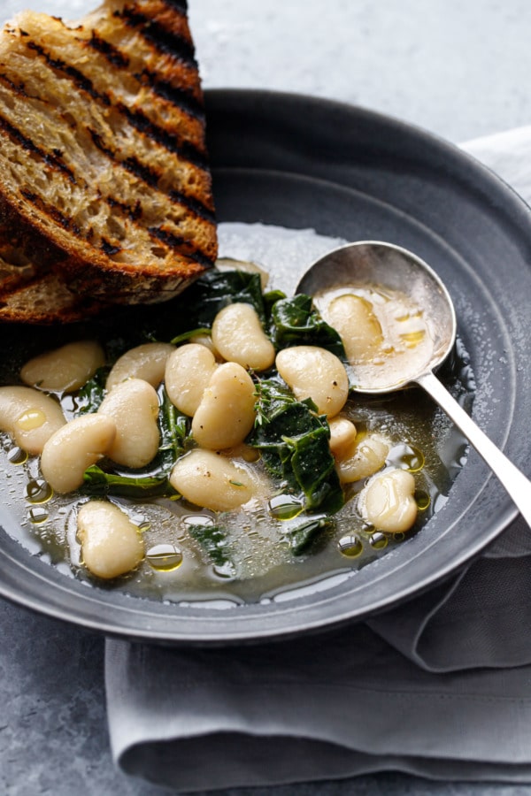 Closeup bowl of cooked white lima beans in broth, with wilted kale and grilled bread on the side