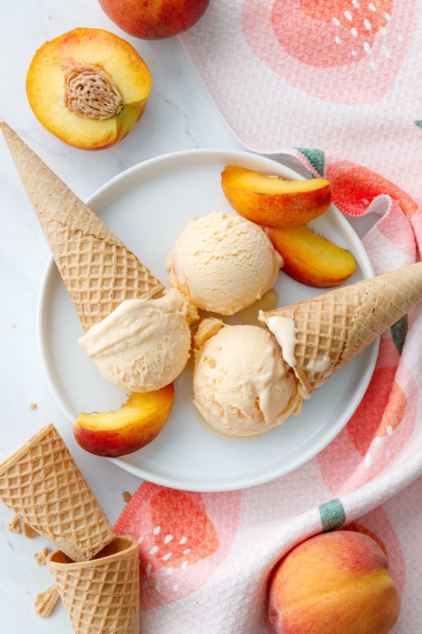 Overhead, three scoops of fresh peach ice cream on a white plate, with peach slices and sugar cones