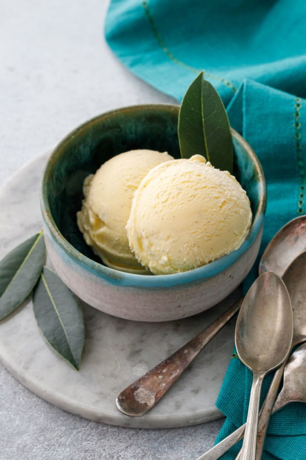 Handmade ceramic bowl with two scoops of vanilla bean bay leaf ice cream, on a marble trivet with spoons and fresh bay leaves