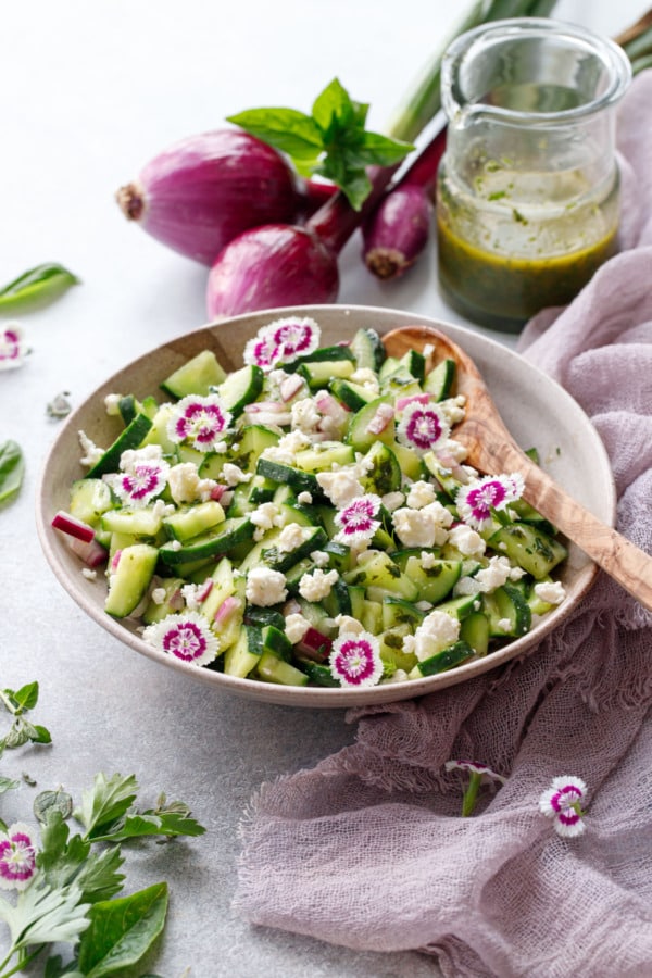 Cucumber salad garnished with edible flowers, in bowl with purple linen and red onions in background.