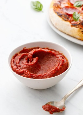 White bowl with homemade pizza sauce and spoon, homemade pizza on a plate in the background