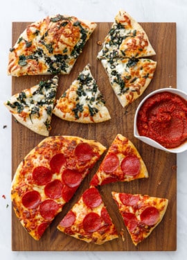 Overhead wooden cutting board with two sliced pizzas (pepperoni and pizza bianca), with a bowl of sauce