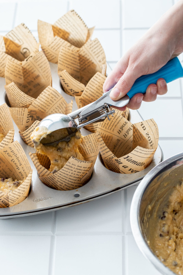 Scooping raw banana bread muffin batter into tulip muffin cups with a blue ice cream scoop