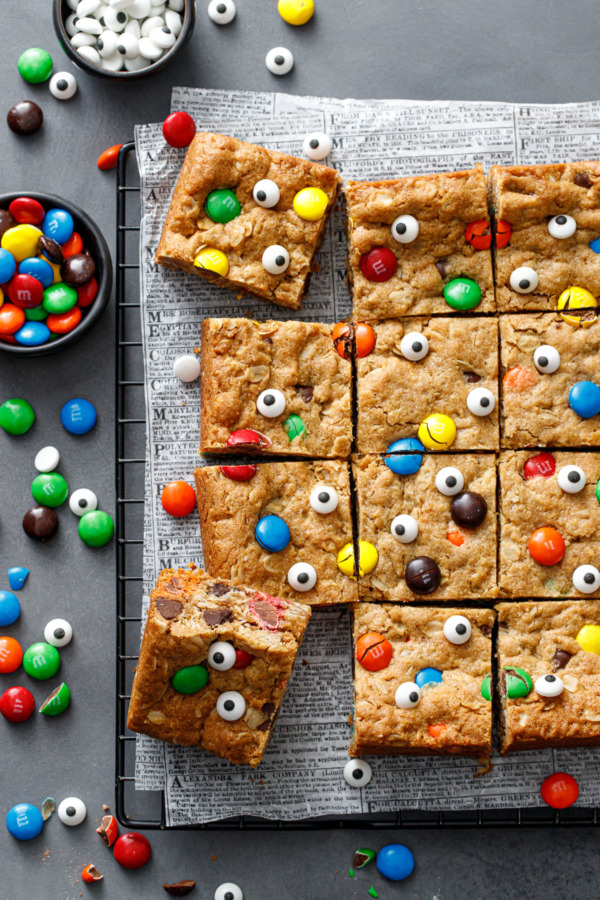 Overhead, squares of Monster Cookie Bars on a dark gray background, small bowl of M&Ms and candy eyeballs
