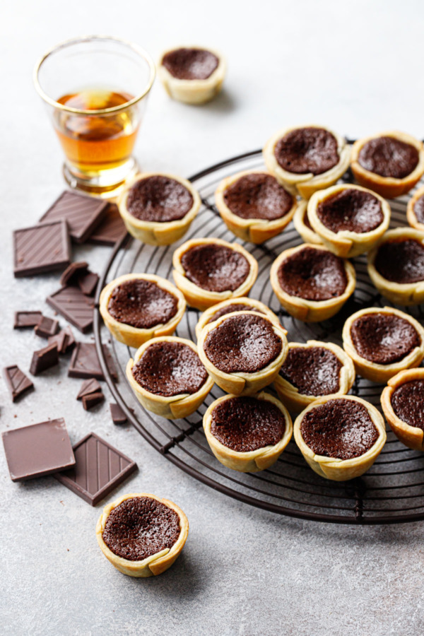 Pile of Mini Bourbon Fudge Pies on a round wire rack, shot of bourbon in the background