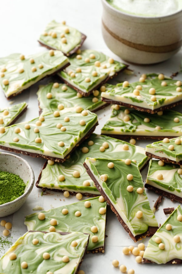 Uneven cut pieces of Matcha White Chocolate Crunch Bark, with small bowl of matcha tea powder and a cup of matcha latte in the background