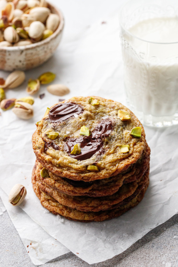 Stack of four Salted Pistachio & Dark Chocolate Chunk Cookies on crinkled parchment, with glass of milk and bowl of shelled pistachios in the background.