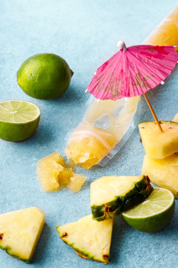 Mai Tai Ice Pops squeezed out of the plastic tube to show the slush-like texture, pieces of pineapple, limes, and a pink cocktail umbrella.