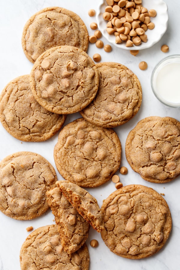 Overhead, scattered Salted Peanut Butter Chip Cookies on marble, with a bowl of pb chips and glass of milk.