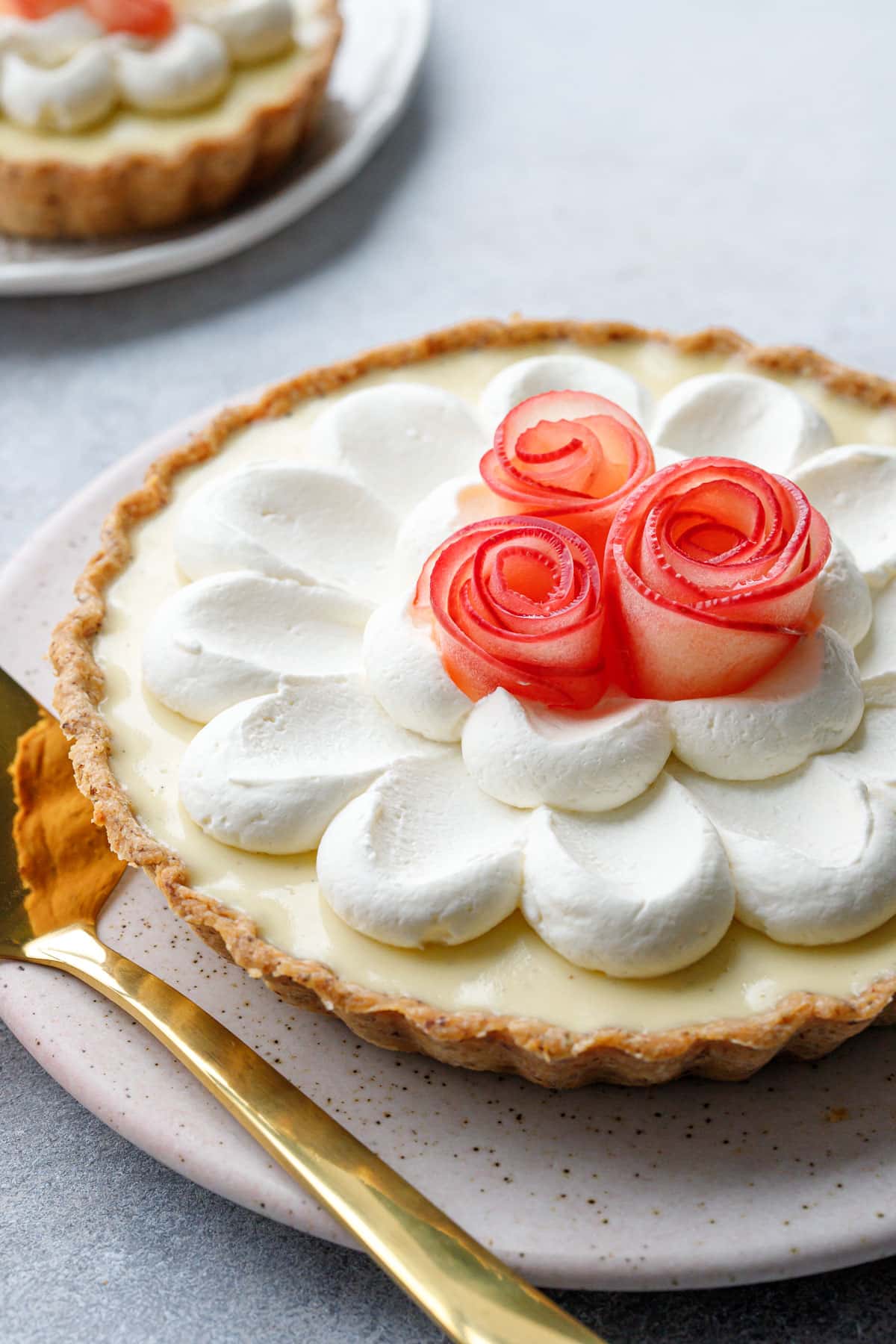 Closeup on decoration of a Caramel Apple Mousse Tart, a flower of piped whipped cream topped with three pink apple roses and a gold cake server on the side.