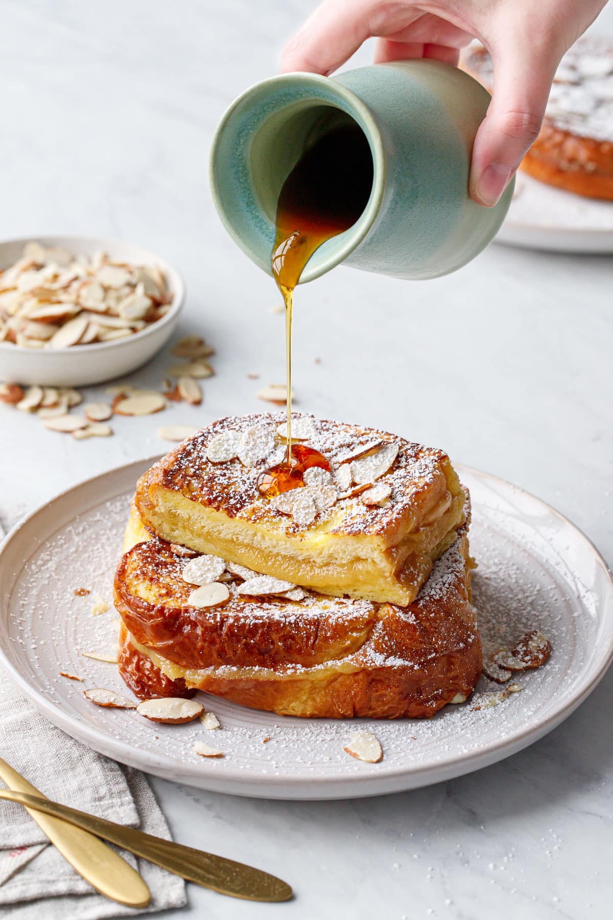 Action shot of drizzling syrup onto a stack of Marzipan-Stuffed French Toast topped with sliced almonds and dusted with powdered sugar.